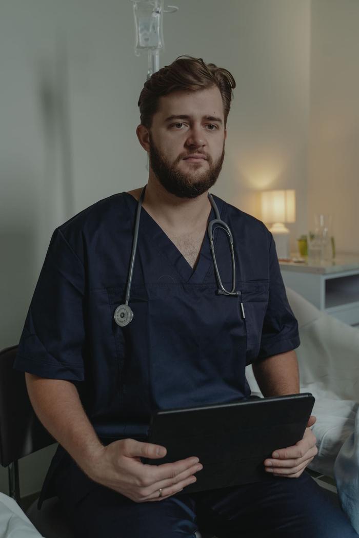 A healthcare worker in navy scrubs seated with a tablet in a patient room.