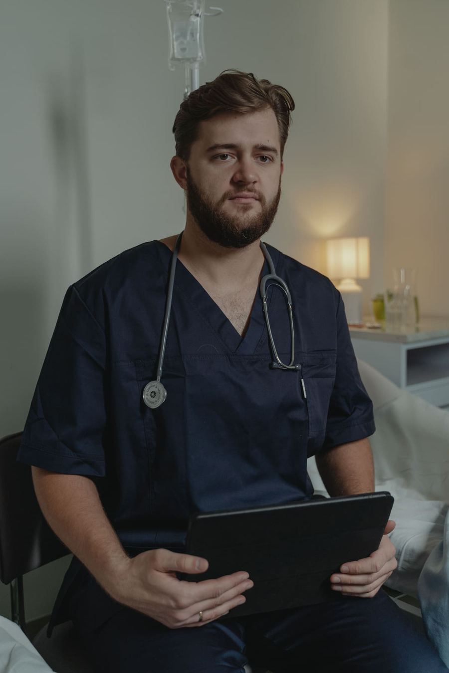 A healthcare worker in navy scrubs seated with a tablet in a patient room.