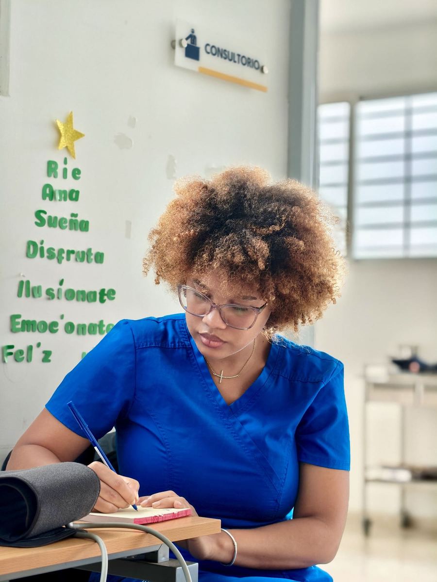 A medical assistant in blue scrubs writing notes at a desk beside a blood pressure cuff.
