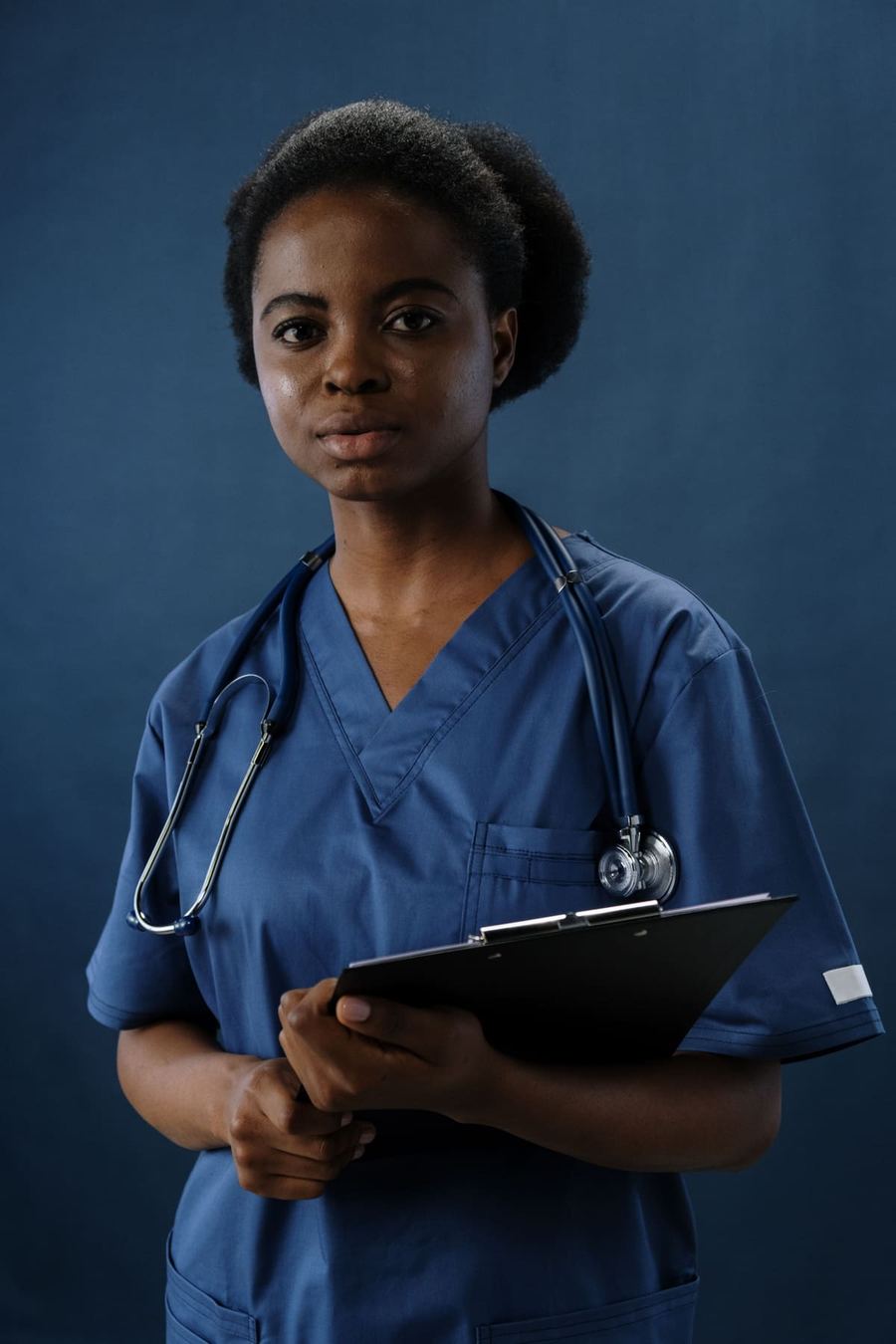 A nurse in blue scrubs holding a clipboard, a stethoscope draped around their shoulders.