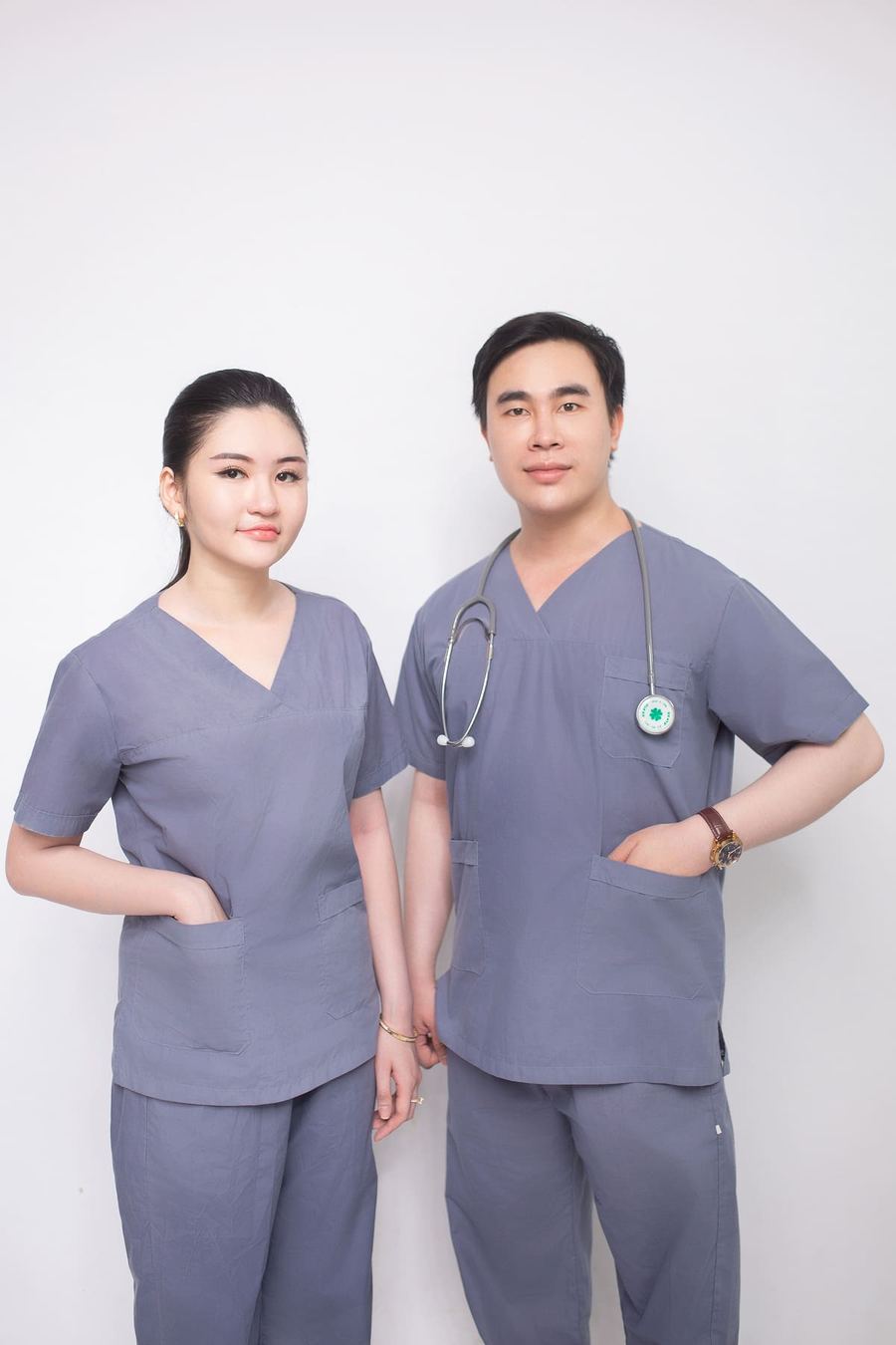 Two healthcare workers in grey surgical scrubs standing against a white wall.